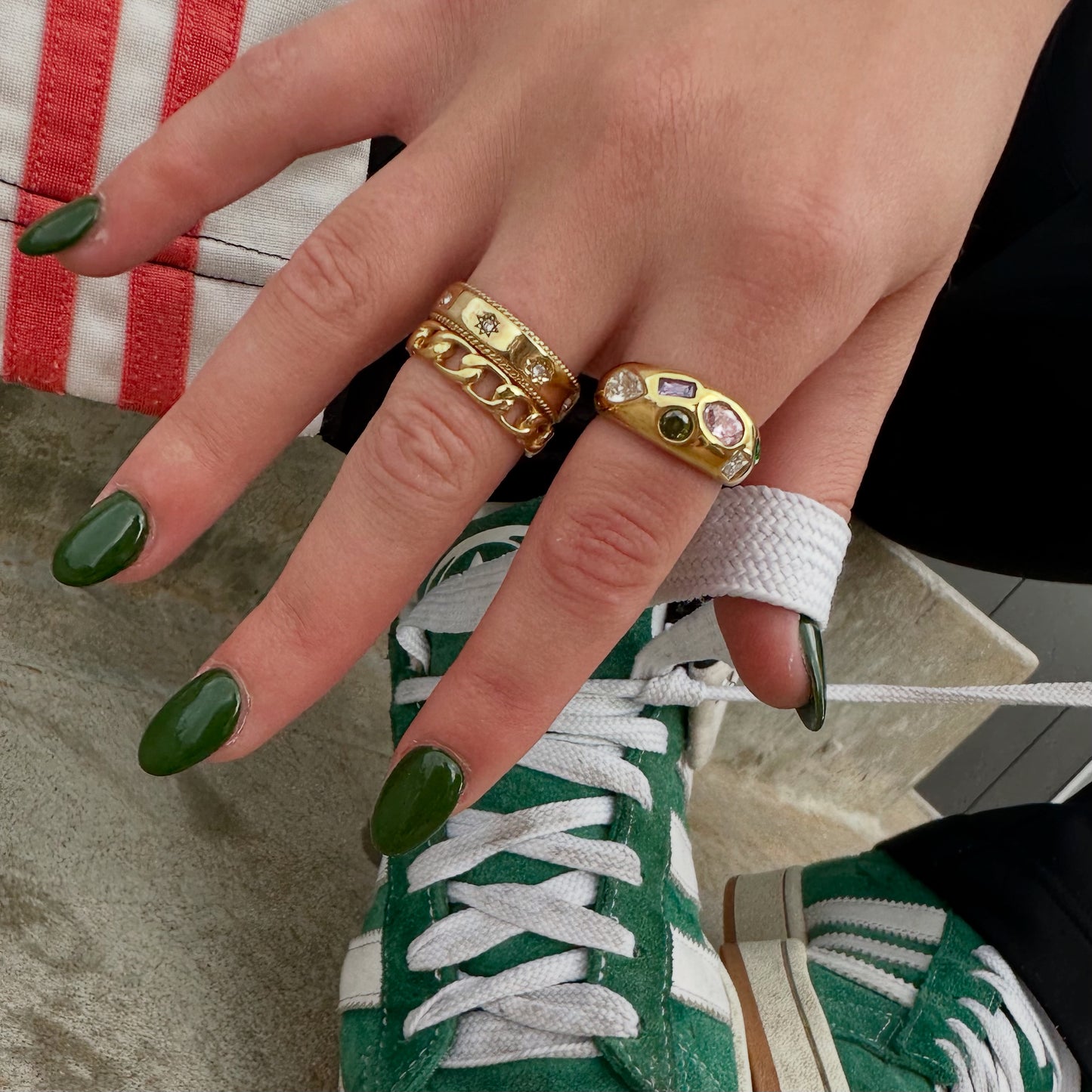 Close-up of a hand with glossy green nail polish tying green-and-white sneakers, styled with three statement gold rings from EVER Jewellery on index and ring fingers.
