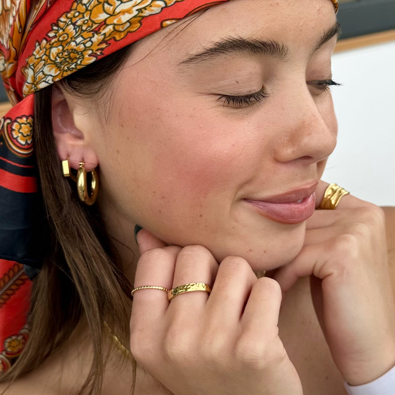 Close-up of a woman in a red headscarf wearing the Medley Ring and gold hoop earrings, smiling with her chin resting on her hand.