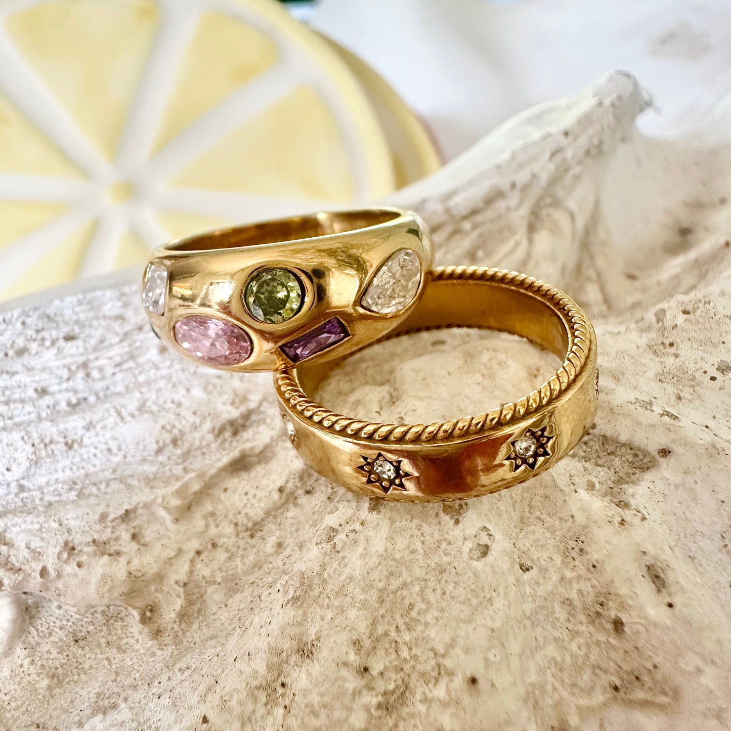 Close-up of two gold EVER rings displayed on a textured seashell—one featuring multicoloured gemstones, the other with engraved star details.