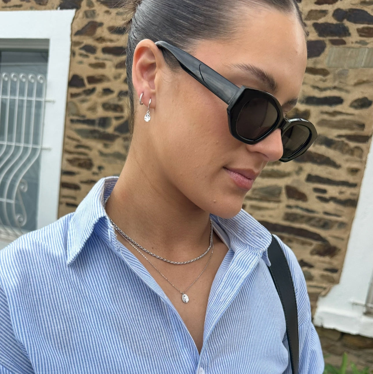 Woman wearing layered Harmony and Perform Silver Necklaces with sunglasses and a blue shirt with a stone wall background