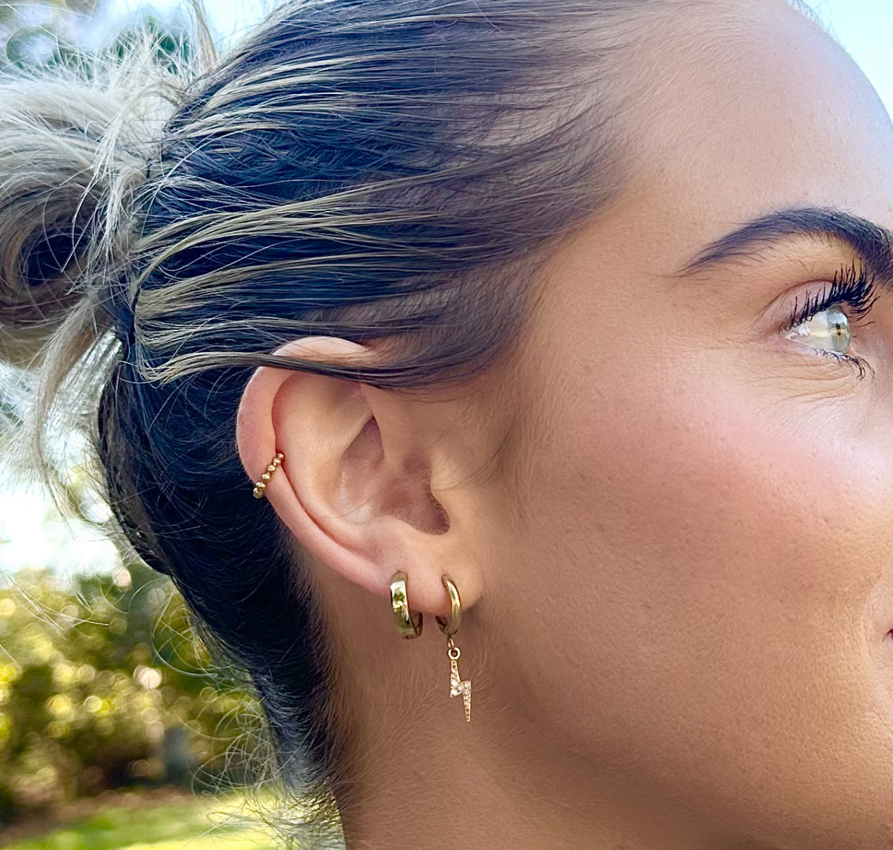 Close-up of a person wearing gold hoop earrings with a blurred outdoor background