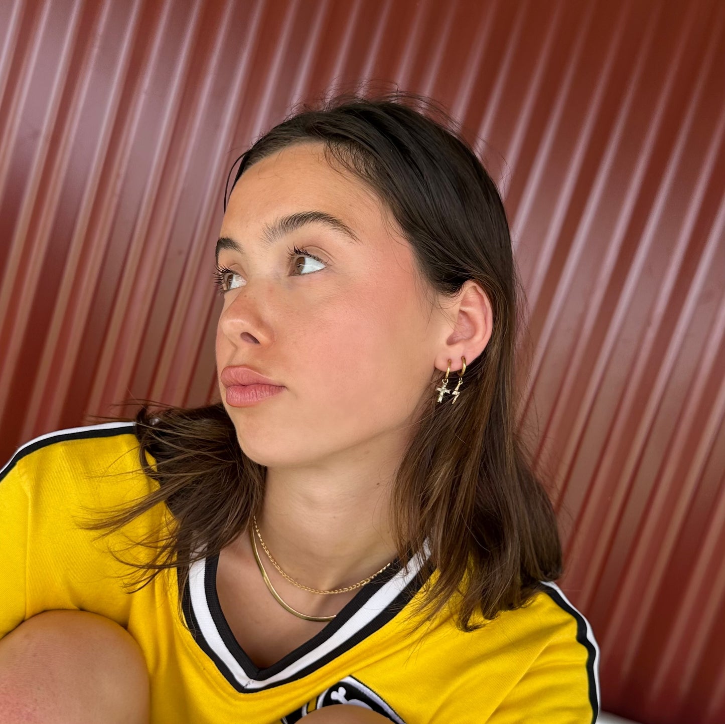 Young woman wearing a yellow sports jersey, styled with stacked gold huggie earrings including lightning bolt charms, seated against a corrugated red metal background.