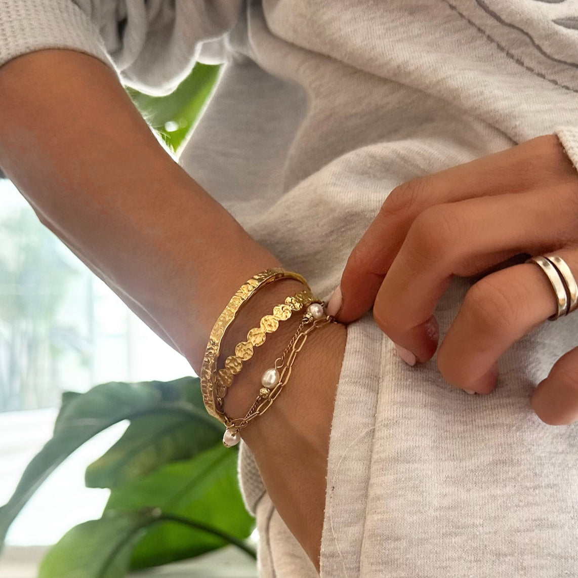 Close-up of the EVER Gold Press Play Cuff stacked with a pearl bracelet and gold ring, worn with a light knit top and hand in pocket.