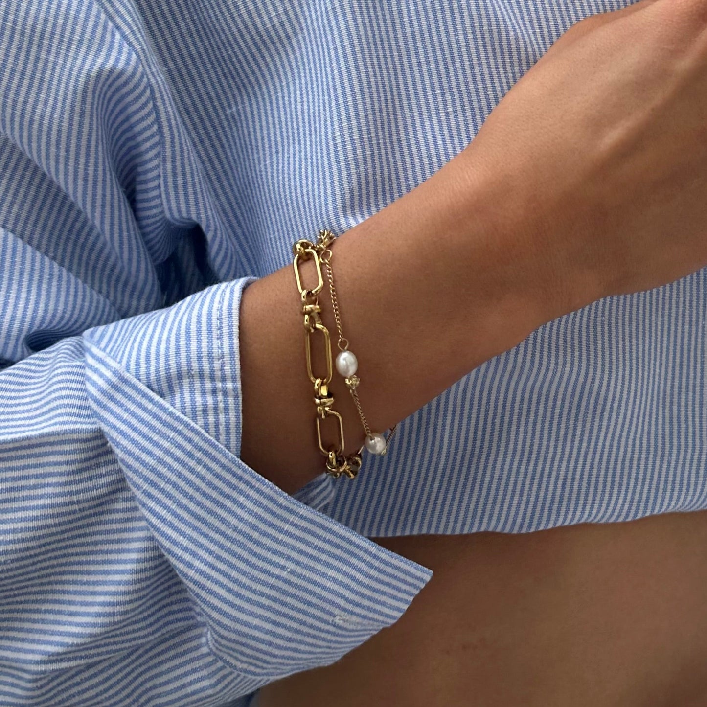 Close-up of a woman wearing the EVER Off Court Pearl Bracelet in gold with freshwater pearls, layered with a chunky gold chain bracelet, pearl pendant necklace, and multiple gold rings, styled with a blue striped shirt and black belt.