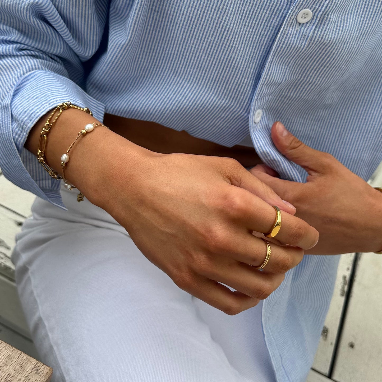 Close-up of a woman wearing the EVER Off Court Pearl Bracelet in gold with freshwater pearls, stacked with a gold chain bracelet and two gold rings, styled with a blue striped shirt and white pants.