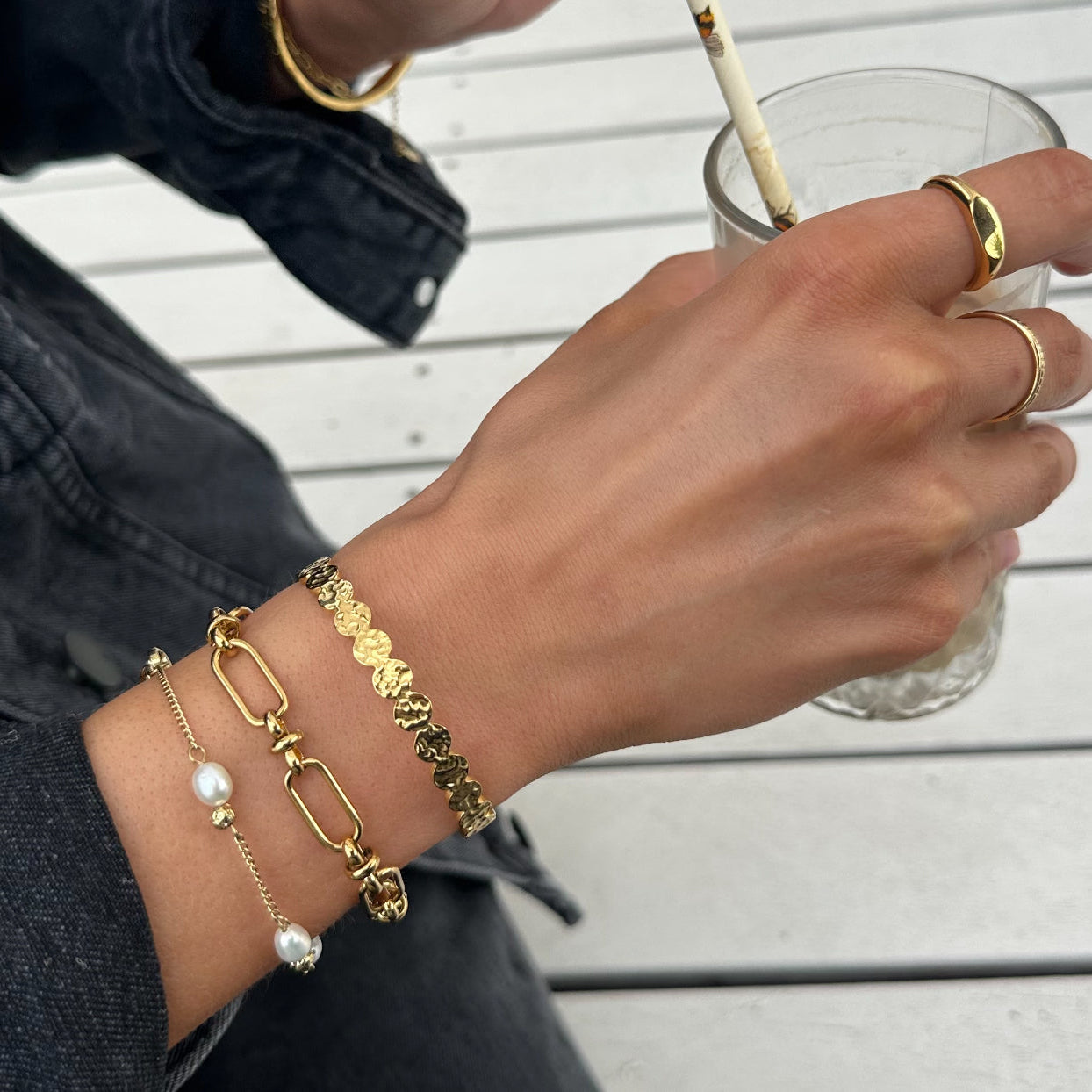 Woman’s hand wearing the EVER Off Court Pearl Bracelet in gold with freshwater pearls, styled with two gold rings and another gold bracelet, holding a glass at an outdoor café.