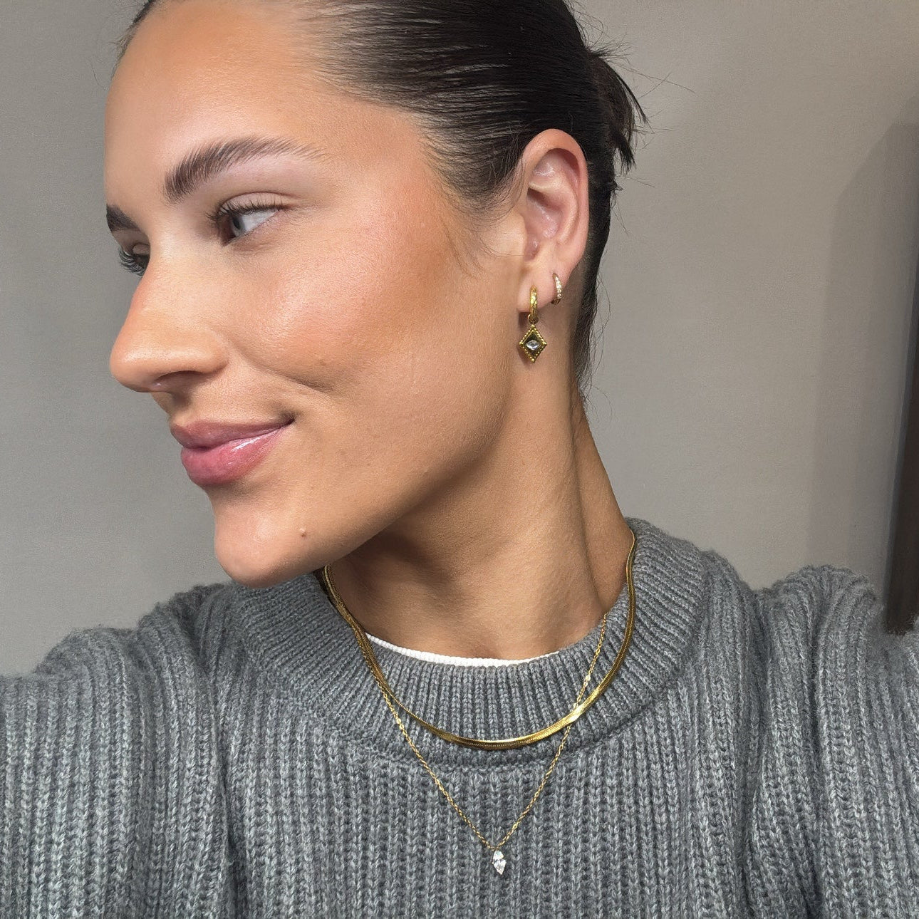 Woman wearing a grey sweater with a neutral background, wearing a stack earrings and stack necklaces