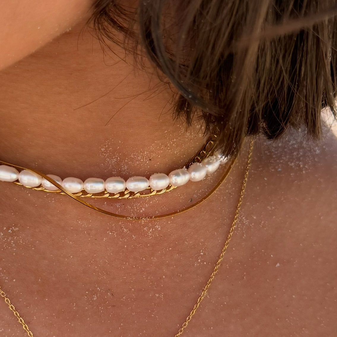 Close-up of a person wearing a pearl necklace and gold chain on a sunlit background