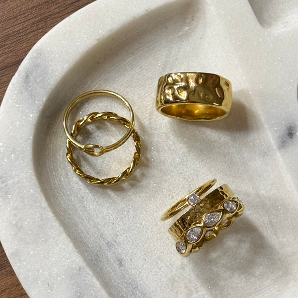 Three gold rings displayed on a white marble tray. One ring is chunky and hammered, one is a delicate twisted band, and one is a stacked ring with clear teardrop gemstones.
