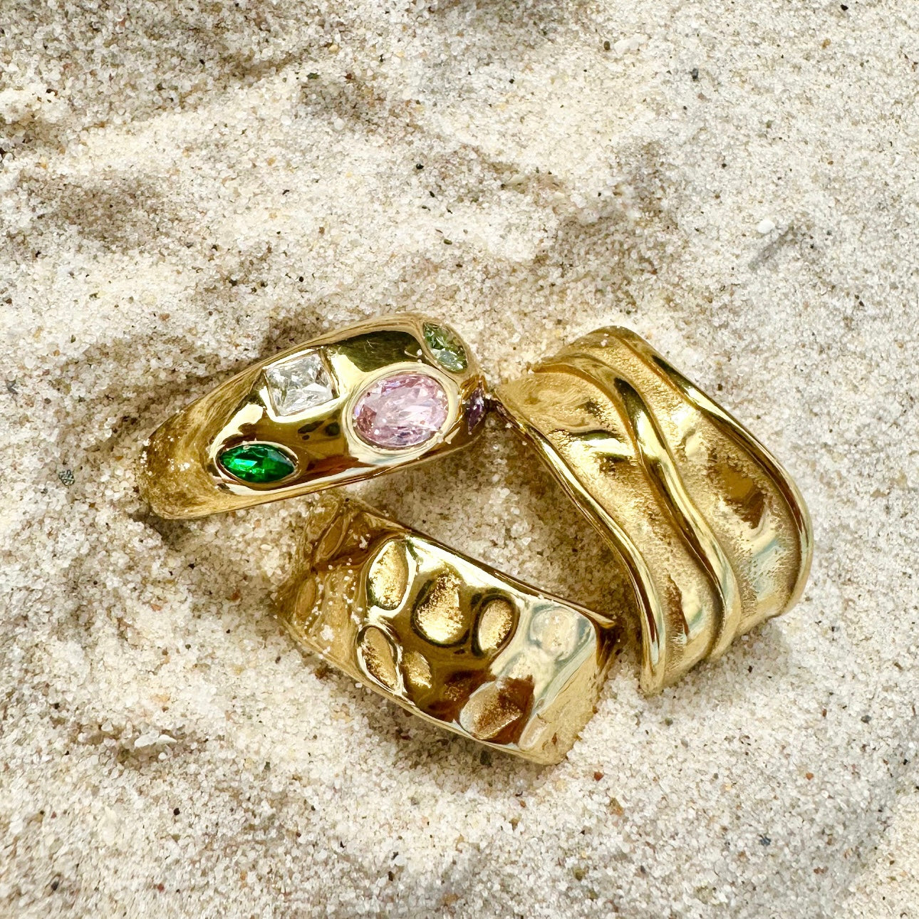 Three EVER gold rings placed on a sandy textured surface, featuring gemstone settings and bold engraved styles.