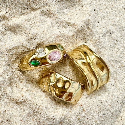 Three EVER gold rings placed on a sandy textured surface, featuring gemstone settings and bold engraved styles.