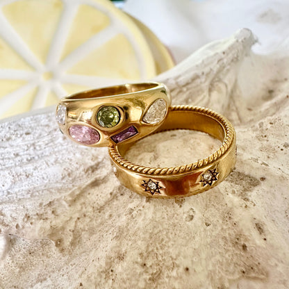 Close-up of two gold EVER rings displayed on a textured seashell—one featuring multicoloured gemstones, the other with engraved star details.