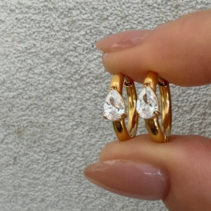 Gold hoop earrings with pear-shaped gemstones held by a hand against a neutral background