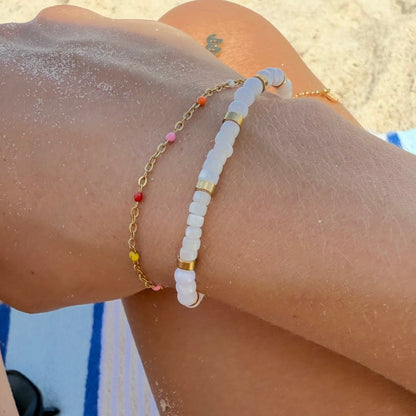 A close-up of a person's wrist at the beach wearing two bracelets: one white beaded bracelet with gold accents and one gold chain bracelet with small multicolor beads.
