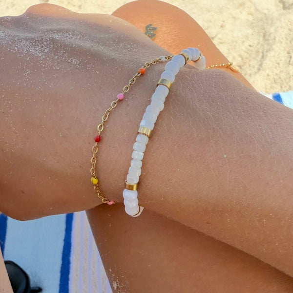 A close-up of a person's wrist at the beach wearing two bracelets: one white beaded bracelet with gold accents and one gold chain bracelet with small multicolor beads.
