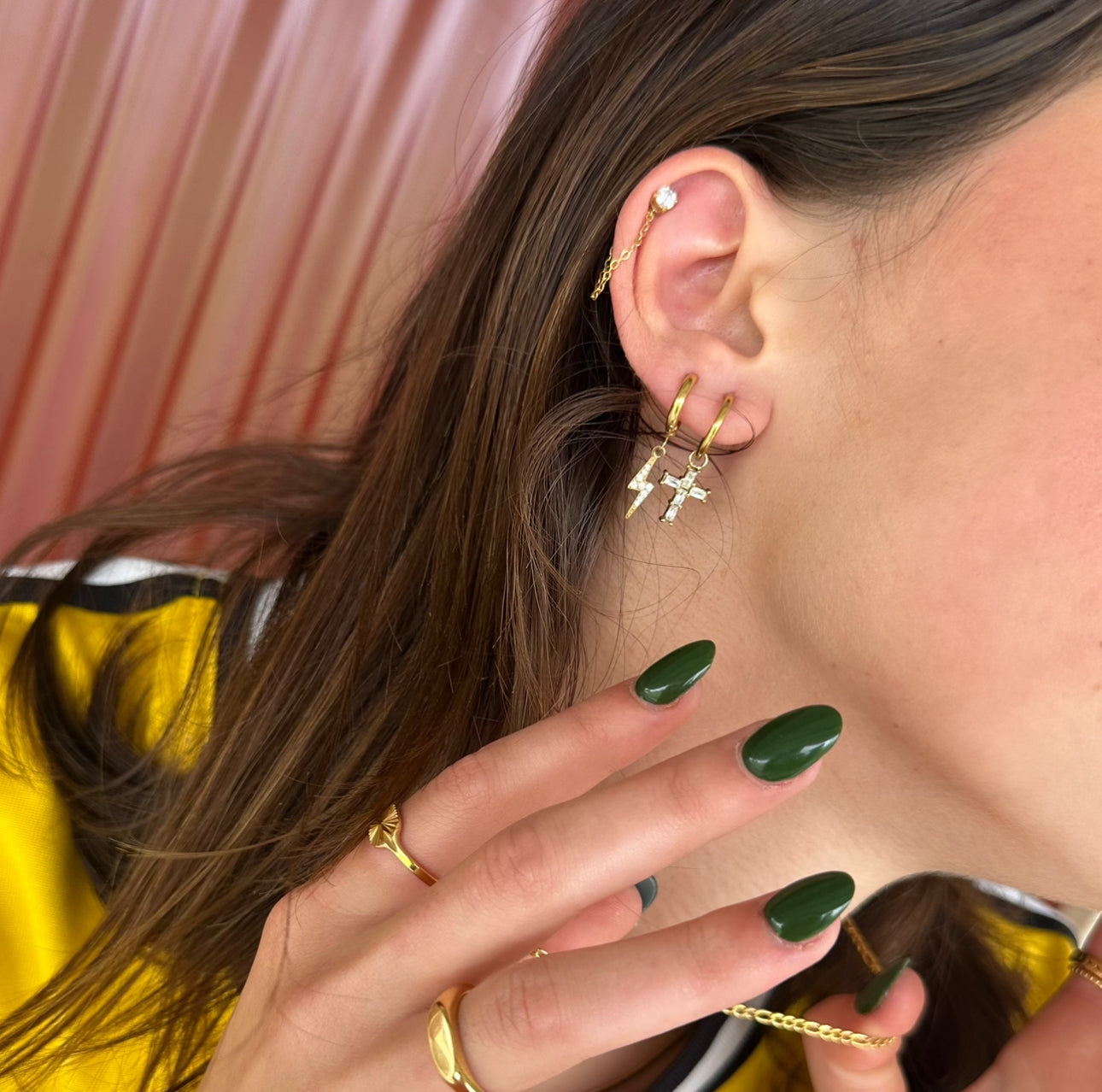 Close-up of a woman's ear wearing EVER's Sprint Huggie and Bolt Huggie Earrings stacked together in gold, styled with layered gold rings and green nails.