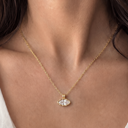 A close-up of a model's neck and chest wearing a delicate gold chain necklace with an oval clear gemstone pendant.