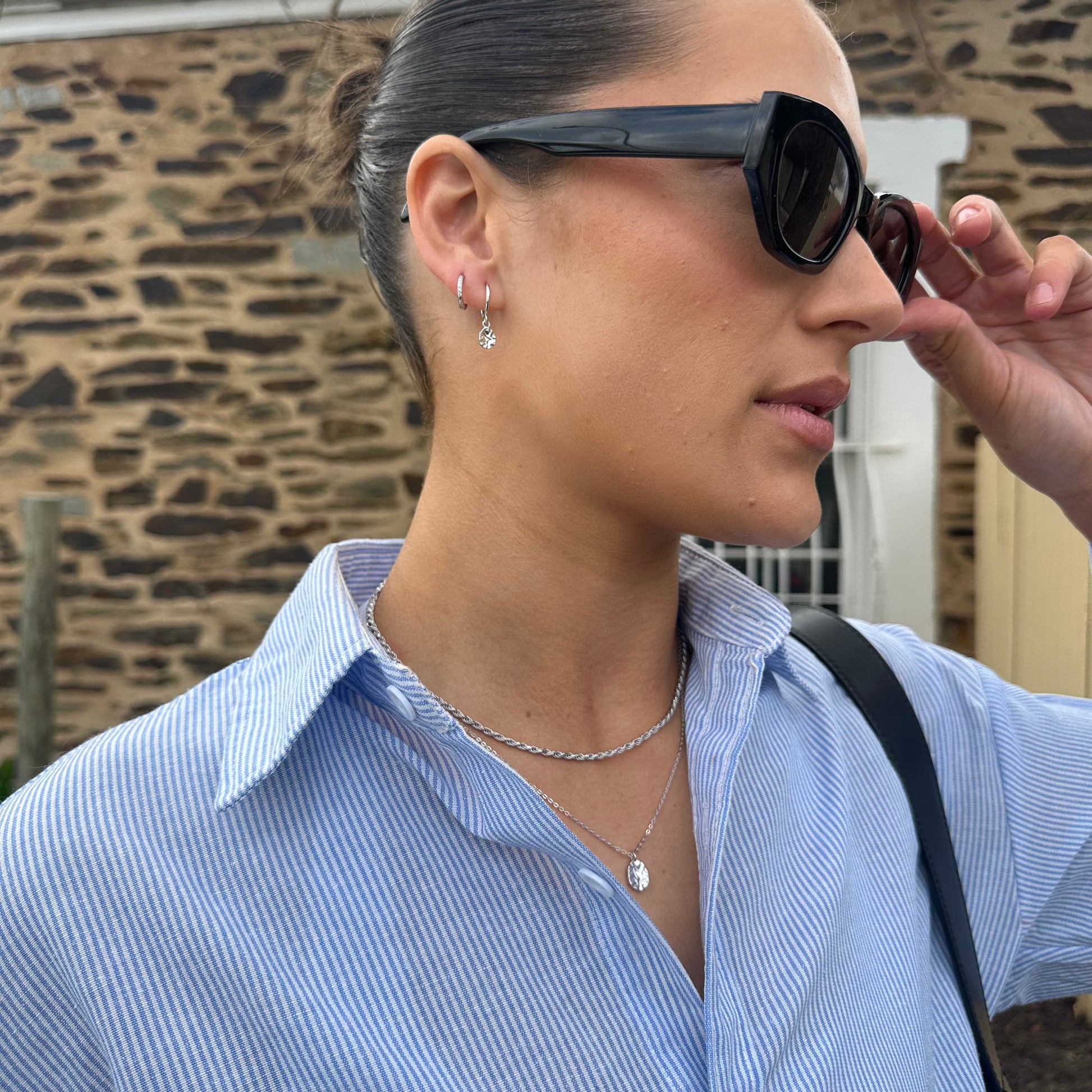 Woman adjusting sunglasses wearing Silver Harmony Earringswith a stone wall background