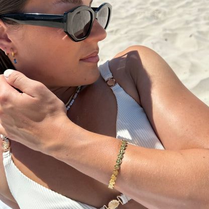 Woman on a beach wearing sunglasses and a white top, with a close-up of her arm and jewelry.