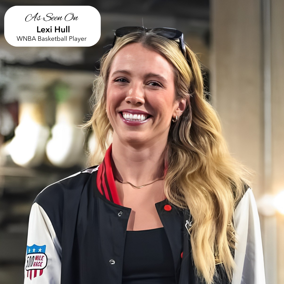 Woman with blonde hair wearing a black and white jacket with a logo, smiling outdoors.