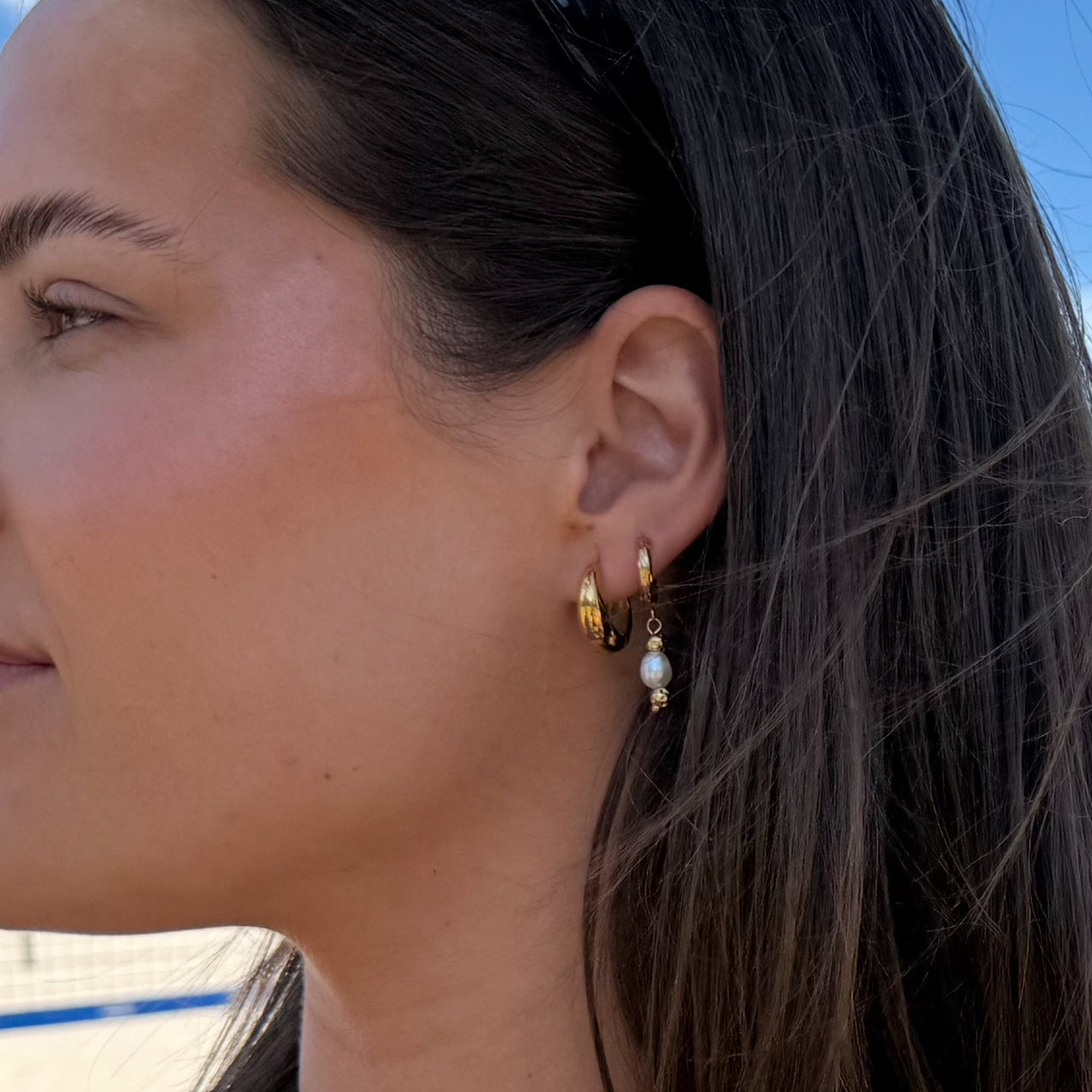 Close-up of a woman wearing gold hoop earrings with pearls against a blue sky.