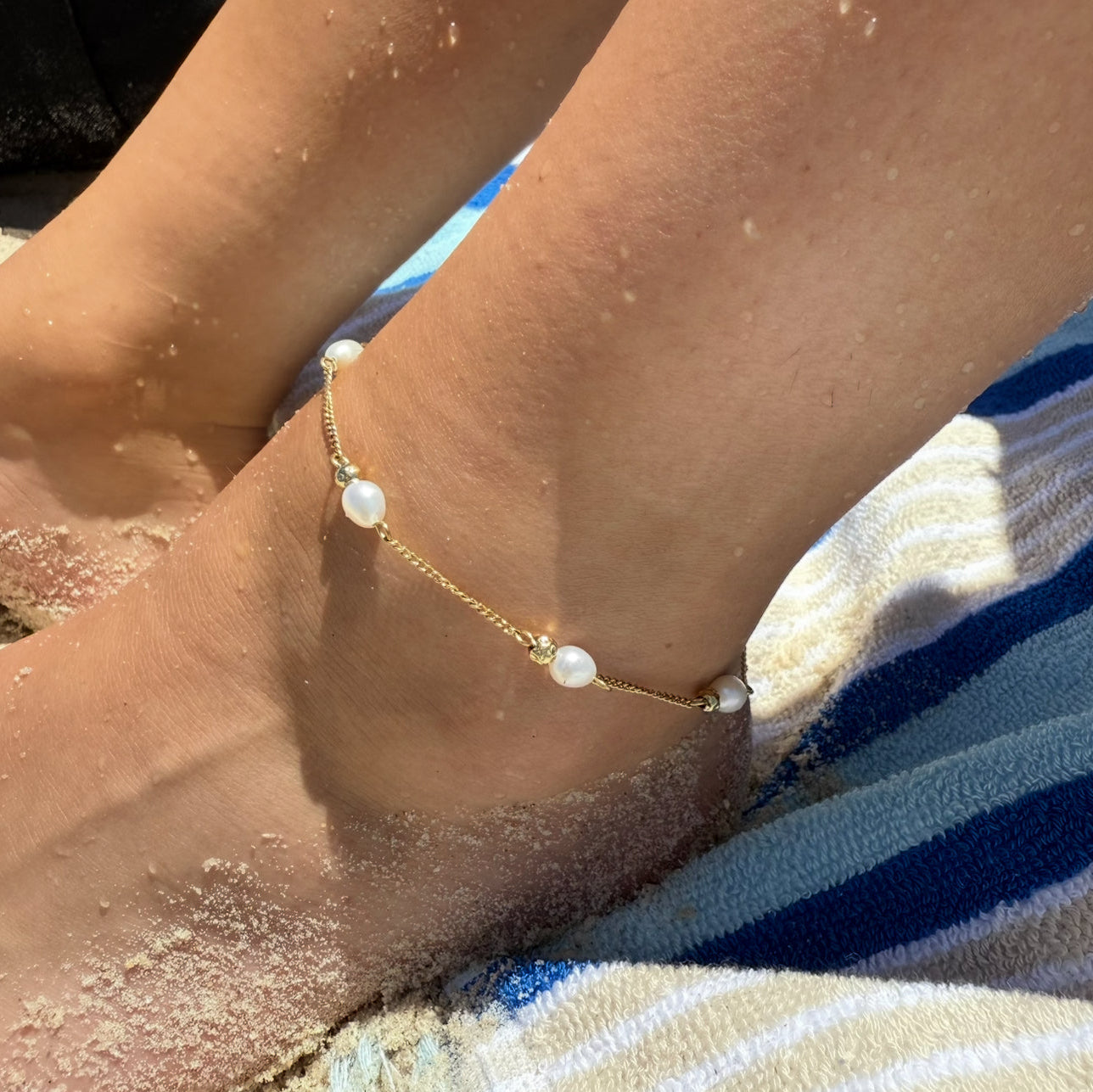 Anchored foot wearing a gold anklet with pearls on a sandy surface.
