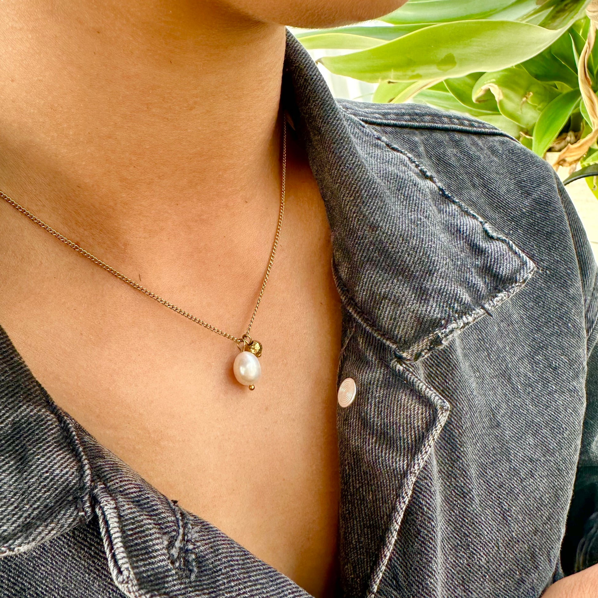 Person wearing the Off Court Pearl Necklace, gold necklace with a pearl pendant, standing outdoors with greenery in the background.