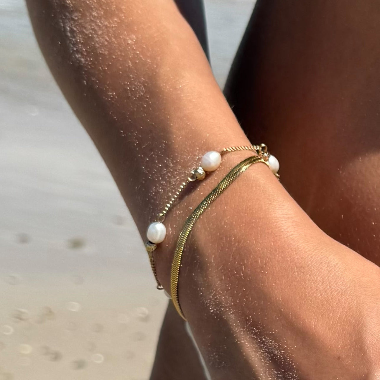 Close-up of a person's arm with gold and pearl bracelet on a beach.