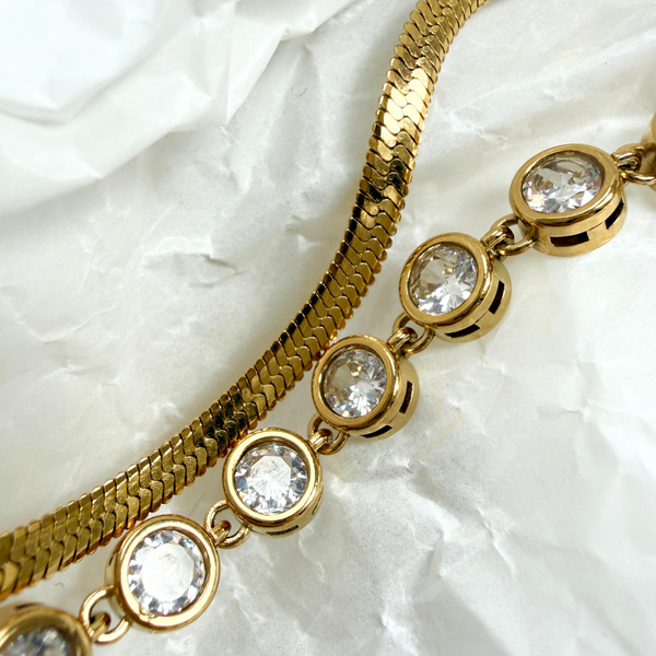 A close-up of a gold-toned chain necklace with oval clear gemstones in bezel settings, laid on a crumpled white fabric background.
