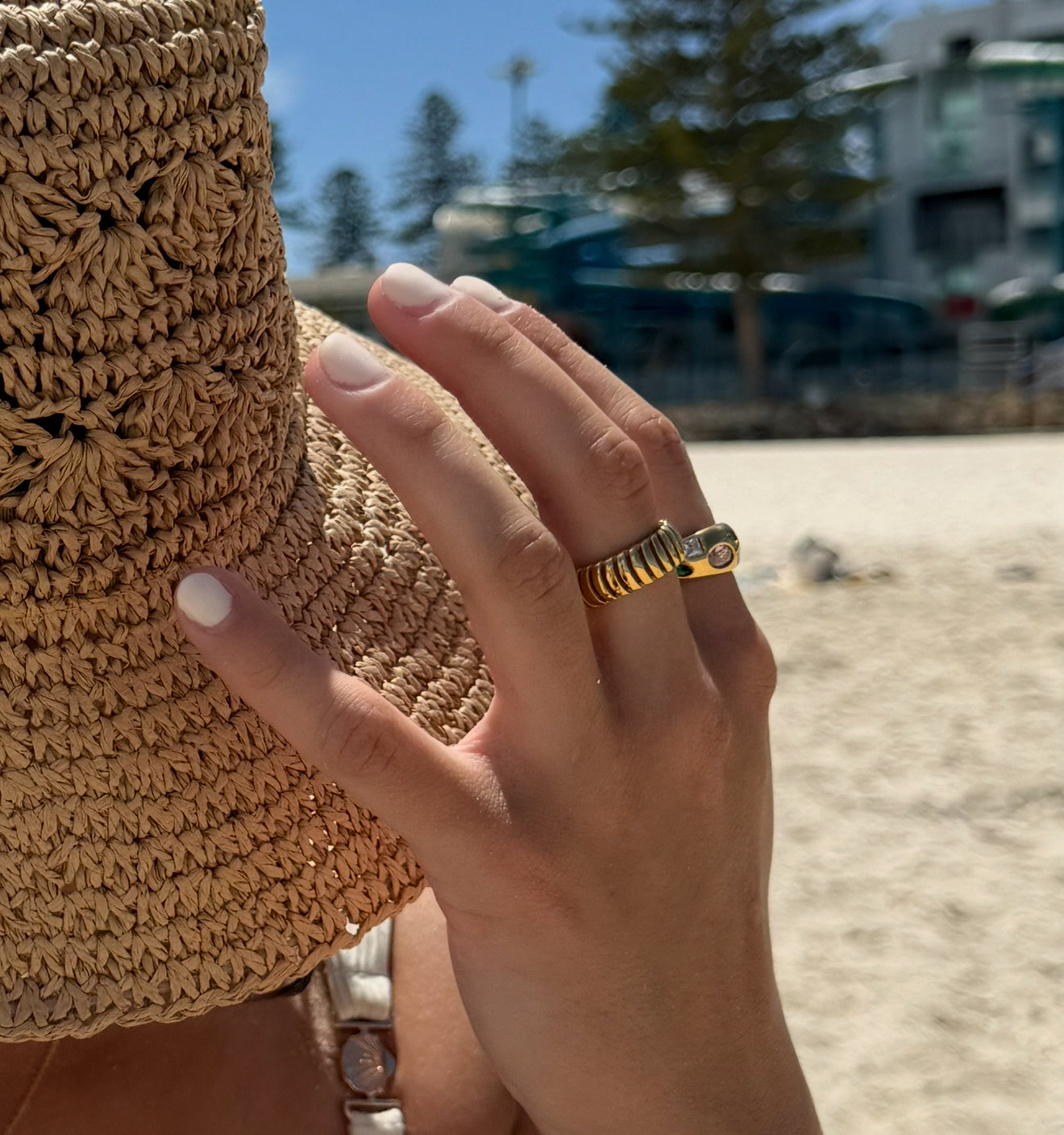 Hand wearing a gold ring with a textured straw hat on a beach.