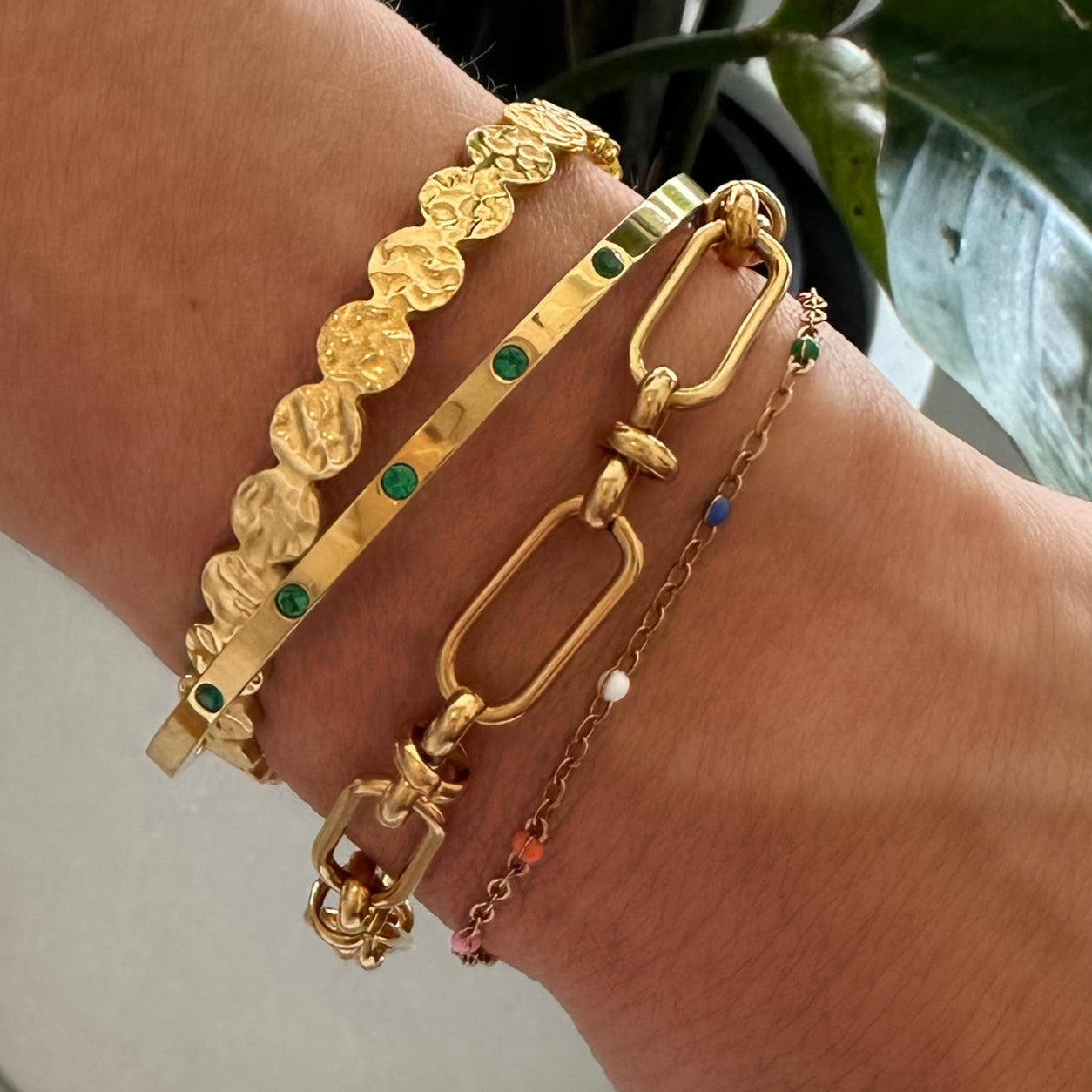 Stacked gold bracelets on a wrist, featuring the EVER Colour Wave Bracelet with multi-coloured beads alongside hammered and emerald-stone gold cuffs.