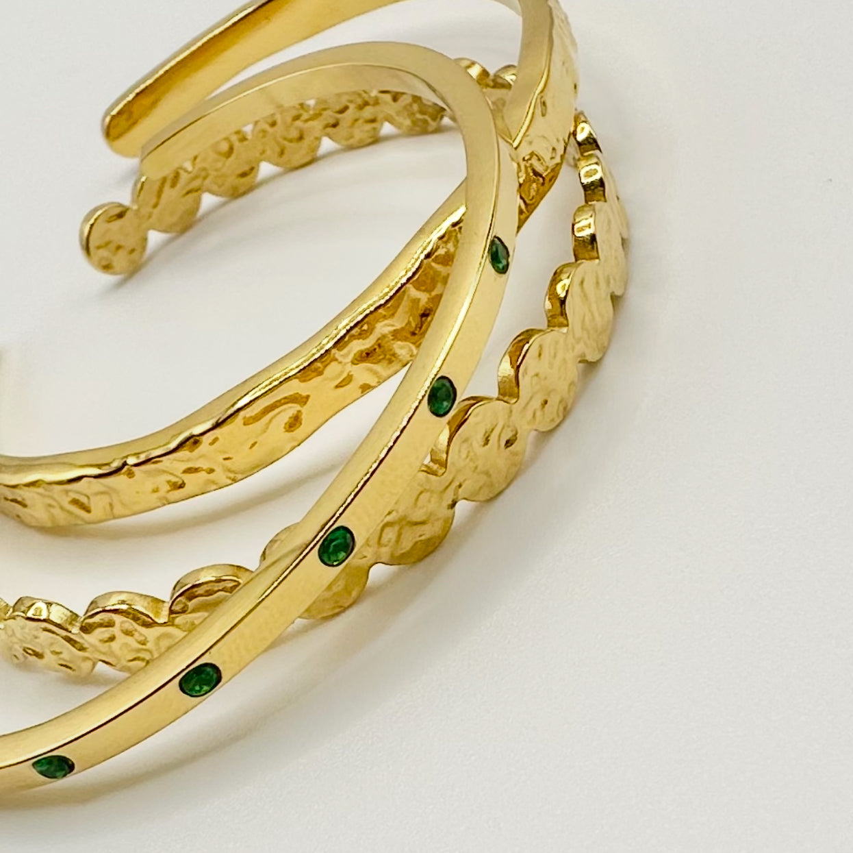 Close-up flatlay of three EVER Jewellery gold hammered cuffs, one accented with emerald green stones, on a clean light background.