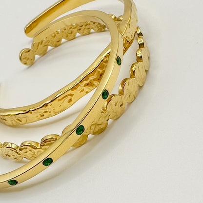 Close-up flatlay of three EVER Jewellery gold hammered cuffs, one accented with emerald green stones, on a clean light background.