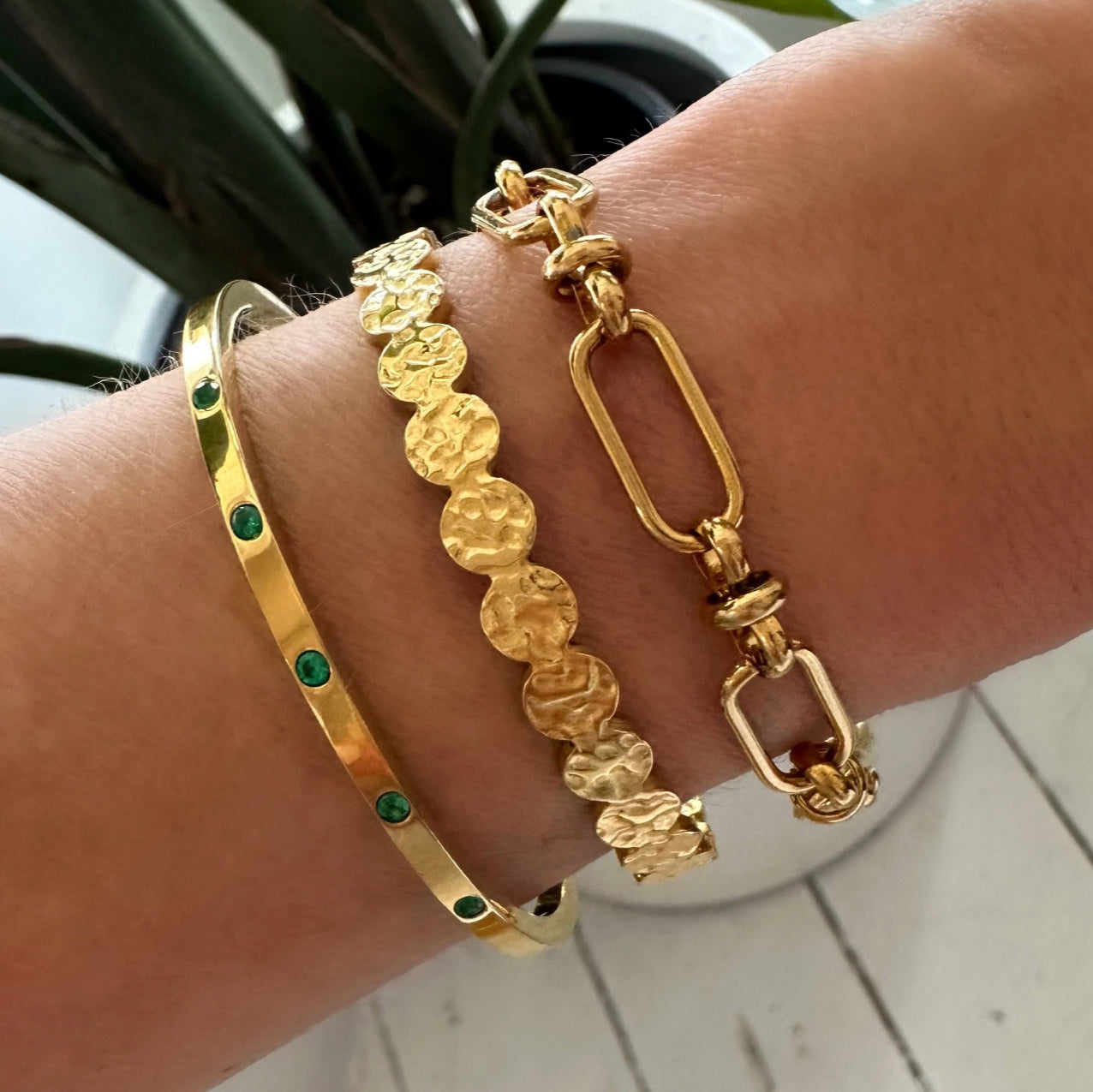 Close-up of a wrist wearing layered EVER Jewellery gold bracelets including the Playmaker Cuff with emerald stones, Signal Bracelet, and Press Play Cuff, photographed indoors with a blurred background.