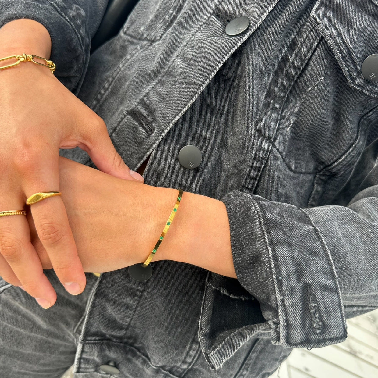 Model wearing EVER Jewellery Playmaker gold cuff bracelet with emerald green stones, styled with a gold ring and black denim jacket.