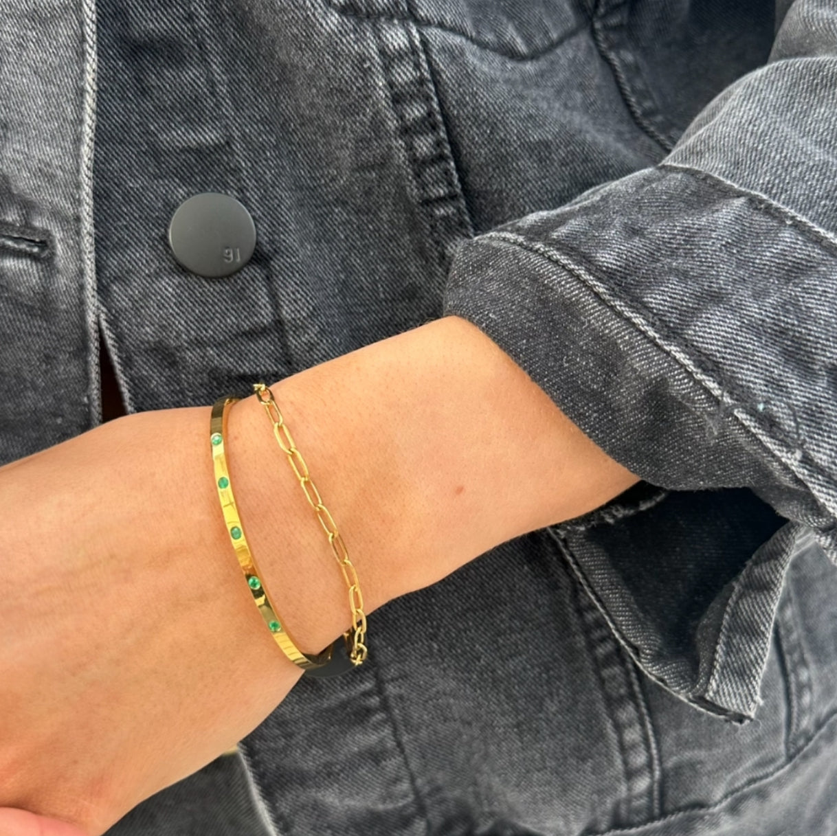 Close-up of EVER Jewellery Playmaker gold cuff bracelet with emerald green stones, stacked with a gold chain bracelet, worn with a black denim jacket.