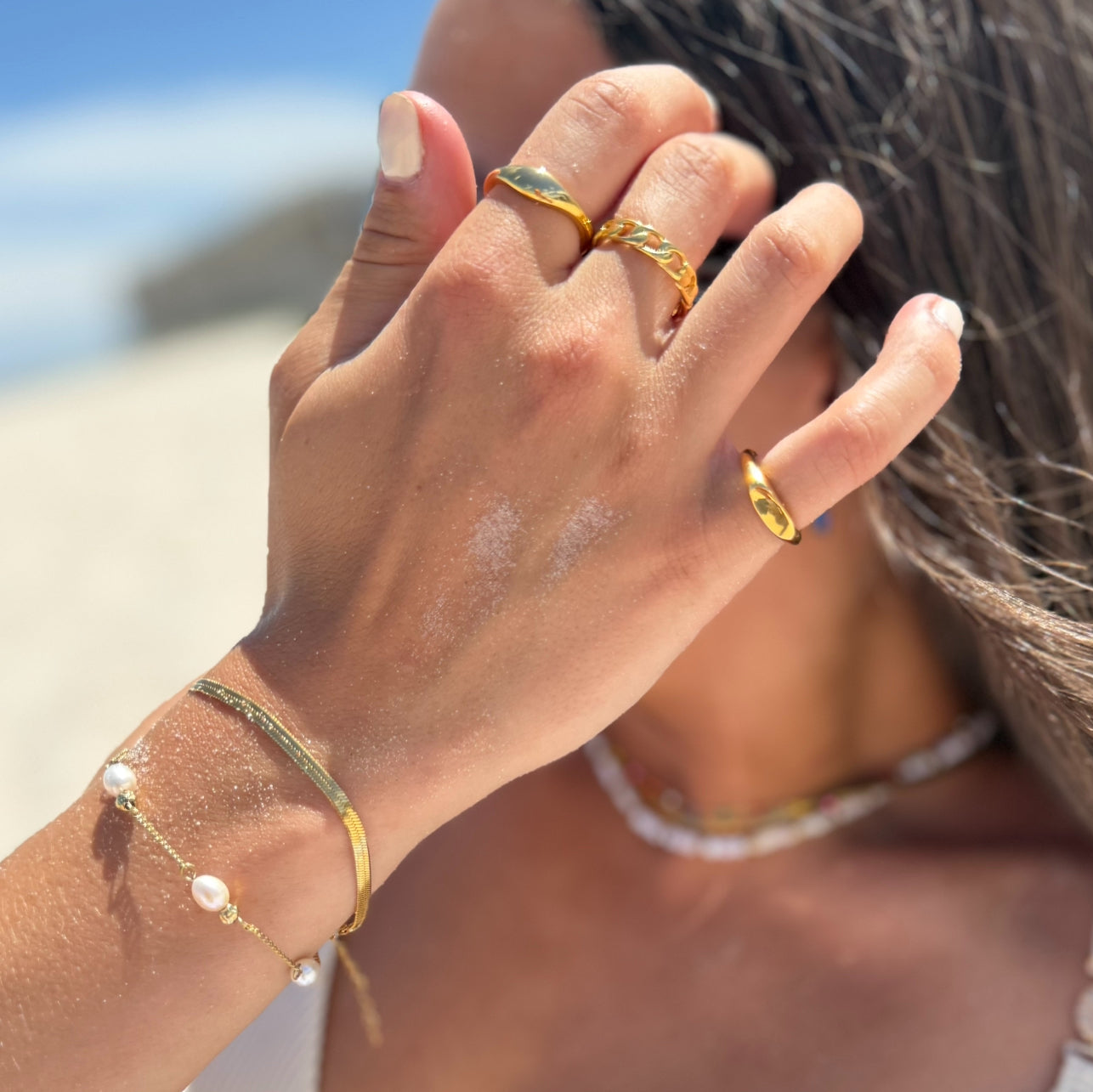 Close-up of a hand wearing gold rings and bracelets with a blurred beach background